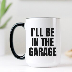 May include: White ceramic coffee mug with a black rim and handle. The mug has black text that reads "I'll be in the garage".