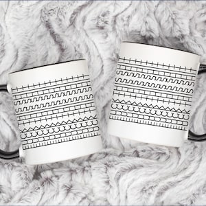 May include: Two white ceramic mugs with black handles and rims. Each mug features a black geometric pattern of lines, squares, triangles, and curves. The mugs are displayed on a fluffy, white, textured surface.
