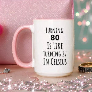 May include: White ceramic coffee mug with a pink rim and handle. The mug has black text that reads "Turning 80 is like turning 27 in Celsius".