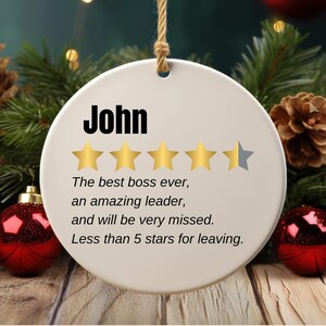 May include: A round, white Christmas ornament with the name "John" in bold black letters. The ornament features four gold stars and one gray star, with text below that reads, "The best boss ever, an amazing leader, and will be very missed. Less than 5 stars for leaving."