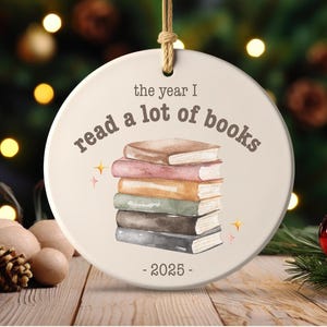 May include: A round, cream-colored ornament with a stack of watercolor books. The text reads "the year I read a lot of books - 2025". The ornament hangs from a natural twine loop.
