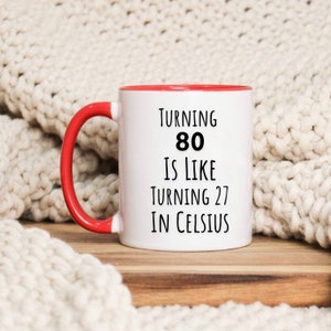 May include: White ceramic mug with a red rim and handle. The mug has black text that reads "Turning 80 is like turning 27 in Celsius."