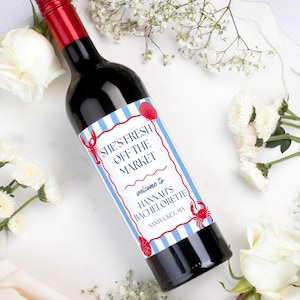 May include: A wine bottle with a red foil cap and a label featuring light blue and white vertical stripes, red lobster and seashell illustrations, and the text: SHE'S FRESH OFF THE MARKET, welcome to HANNAH'S BACHELORETTE, NANTUCKET, MA.