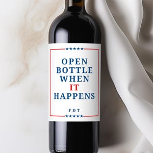 A red wine bottle against a white marble background and white cloth on the right. The wine bottle has a custom wine label that is white with a red border and blue stars on the top and bottom of the border. The text inside the box reads "Open Bottle When It Happens" and smaller "FDT" font on the bottom. Perfect anti-Trump gifts