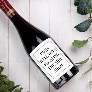May include: A dark glass wine bottle with a red screw cap and a white rectangular label. The label features a black border and the black text: PAIRS WELL WITH ESCAPING THE SHIT SHOW. The bottle is positioned diagonally across the frame.