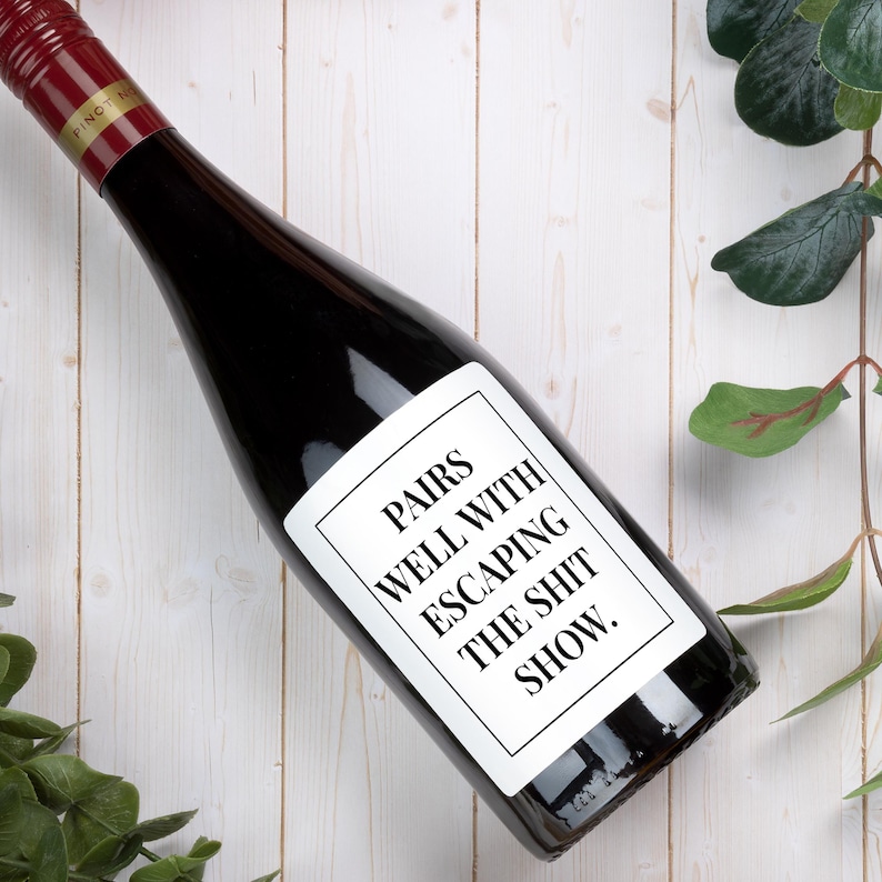 May include: A dark glass wine bottle with a red screw cap and a white rectangular label. The label features a black border and the black text: PAIRS WELL WITH ESCAPING THE SHIT SHOW. The bottle is positioned diagonally across the frame.