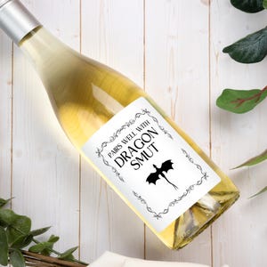 May include: A clear glass wine bottle filled with pale yellow liquid, featuring a white rectangular label with a black decorative border. The label text reads PAIRS WELL WITH DRAGON SMUT, accompanied by a black silhouette of a flying dragon.
