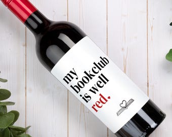 May include: A dark glass wine bottle with a red screw cap and a white label. The label features black text that reads "my bookclub is well red." with the word "red" in red ink, followed by a small red heart and a black line drawing of an open book.