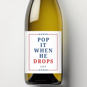 Puede incluir: Una botella de vino con una etiqueta blanca con el texto "POP IT WHEN HE DROPS" en azul y rojo. La etiqueta está enmarcada con un borde rojo y estrellas azules. La botella contiene un líquido amarillo claro.