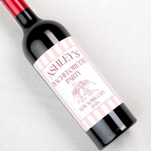 May include: A dark glass wine bottle with a red cap and a custom label featuring vertical light pink and white stripes. The label text reads ASHLEY'S BACHELORETTE PARTY, NEW YORK CITY 2026, accompanied by a line drawing of two clinking cocktail glasses.