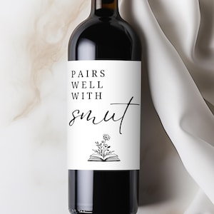 May include: A dark glass wine bottle with a white label. The label reads "PAIRS WELL WITH smut" in black script. Below the text is a small illustration of an open book with flowers.