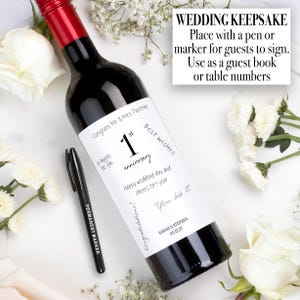 May include: A wine bottle with a red foil cap and a white label featuring the text 1st anniversary, Congrats Mr & Mrs. Fletcher, Best wishes, and Sarah & Stephen 03.12.27. A black permanent marker is positioned next to the bottle for guest signatures.