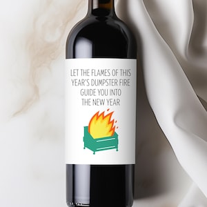 May include: A wine bottle with a label featuring the text "Let the flames of this year's dumpster fire guide you into the new year." The label includes a graphic of a dumpster on fire. The bottle is filled with red wine.