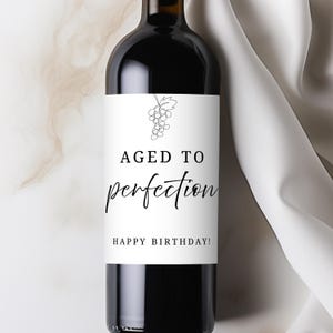 Puede incluir: Una botella de vino tinto con una etiqueta blanca. La etiqueta presenta un dibujo lineal de uvas sobre las palabras "AGED TO perfection" y "HAPPY BIRTHAY!" en una fuente de escritura. La botella está sobre un fondo blanco.