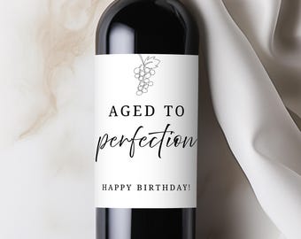 May include: A dark glass wine bottle with a white rectangular label featuring a black line drawing of a grape cluster. The text reads AGED TO perfection and HAPPY BIRTHDAY! in a combination of bold serif and elegant script black typography.