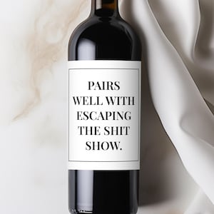 May include: A dark glass wine bottle with a white label. The label reads "PAIRS WELL WITH ESCAPING THE SHIT SHOW." The bottle is set against a neutral background with a white fabric draped on the right side.