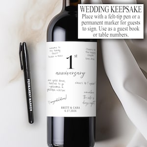 Può includere: Una bottiglia di vino nera con un'etichetta che recita "1st Anniversary". L'etichetta presenta messaggi scritti a mano. Un pennarello indelebile nero è accanto alla bottiglia. È visibile anche il testo "Wedding Keepsake".