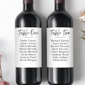 May include: Two dark wine bottles with labels. The labels read "Table One" and "Table Two" in elegant script. Each label lists names, ideal for wedding or event table assignments. The bottles have a dark purple top.