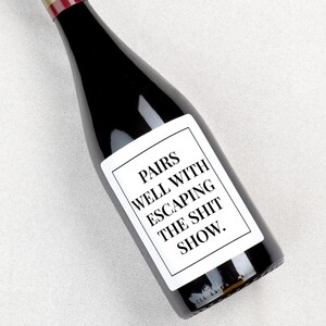 May include: A dark glass wine bottle featuring a rectangular white label with a black border. The label displays the black text: PAIRS WELL WITH ESCAPING THE SHIT SHOW. The bottle has a narrow neck and a red foil seal at the very top of the bottle.