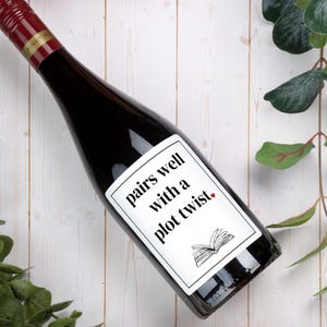 May include: A dark glass wine bottle with a red screw cap and a white rectangular label. The label features the black text "pairs well with a plot twist." followed by a small red heart and a black line drawing of an open book at the bottom.