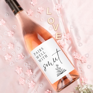 May include: A glass wine bottle filled with pale pink rosé wine, featuring a white rectangular label with black text that reads PAIRS WELL WITH smut and a small illustration of a flower growing out of an open book, set against a pink fabric.