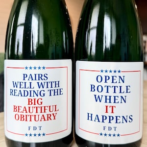 Puede incluir: Dos botellas de vino de vidrio verde oscuro con etiquetas rectangulares blancas. La etiqueta izquierda dice PAIRS WELL WITH READING THE BIG BEAUTIFUL OBITUARY y FDT. La etiqueta derecha dice OPEN BOTTLE WHEN IT HAPPENS y FDT.