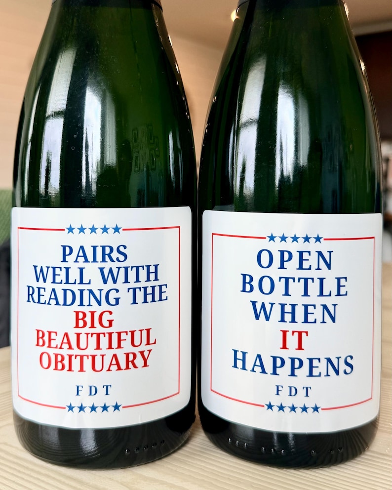 Puede incluir: Dos botellas de vino de vidrio verde oscuro con etiquetas rectangulares blancas. La etiqueta izquierda dice PAIRS WELL WITH READING THE BIG BEAUTIFUL OBITUARY y FDT. La etiqueta derecha dice OPEN BOTTLE WHEN IT HAPPENS y FDT.