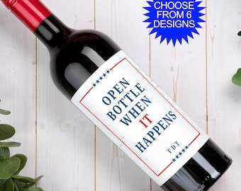 May include: A dark glass wine bottle with a red screw cap and a white rectangular label. The label features a red border, blue stars, and the text "OPEN BOTTLE WHEN IT HAPPENS" in blue and red, with "FDT" below. A blue starburst says "CHOOSE FROM 6 DESIGNS".