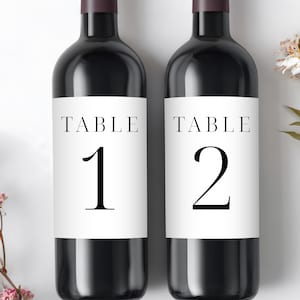 Könnte beinhalten: Zwei dunkle Weinflaschen mit weißen Etiketten. Auf den Etiketten steht "TABLE" über den Zahlen "1" und "2" in fetter, schwarzer Schrift. Die Flaschen haben einen dunkelburgunderfarbenen Deckel und sind vor weißem Hintergrund platziert.