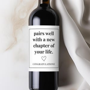 May include: A dark wine bottle with a white label. The label reads "pairs well with a new chapter of your life." with a heart symbol and the word "CONGRATULATIONS!". The bottle is set against a neutral background with a white fabric.