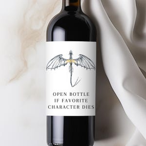 May include: A dark wine bottle with a white label featuring a dragon illustration with gold accents. The text on the label reads "OPEN BOTTLE IF FAVORITE CHARACTER DIES".