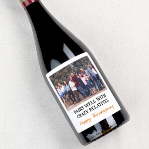 May include: A dark glass wine bottle featuring a rectangular white label with a photograph of a group of people standing outdoors. The label includes the black text PAIRS WELL WITH CRAZY RELATIVES and the orange text Happy Thanksgiving.