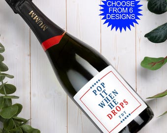 May include: A dark glass Mumm champagne bottle with a custom white rectangular label featuring a red border and blue stars. The text reads POP IT WHEN HE DROPS FDT. A blue starburst graphic in the corner states CHOOSE FROM 6 DESIGNS.