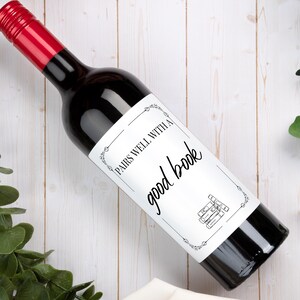 May include: A dark glass wine bottle with a red screw cap and a white rectangular label. The label features a decorative black border, a small illustration of stacked books, and the black text: PAIRS WELL WITH A good book.