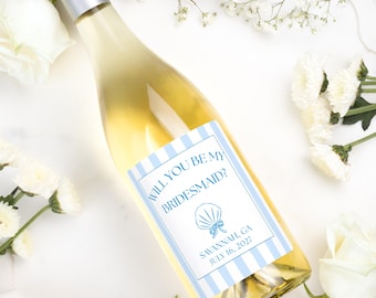 May include: A clear glass wine bottle with a silver cap featuring a rectangular label with light blue vertical stripes. The label text reads Will You Be My Bridesmaid? with a blue seashell graphic and the text Savannah, GA July 16, 2027.