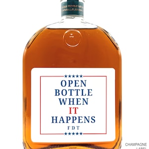May include: A clear, square-shaped bottle filled with amber liquid. The bottle has a white label with the text "OPEN BOTTLE WHEN IT HAPPENS" in blue and red, with stars at the top and bottom. The words "CHAMPAGNE LABEL PICTURED" are at the bottom right.