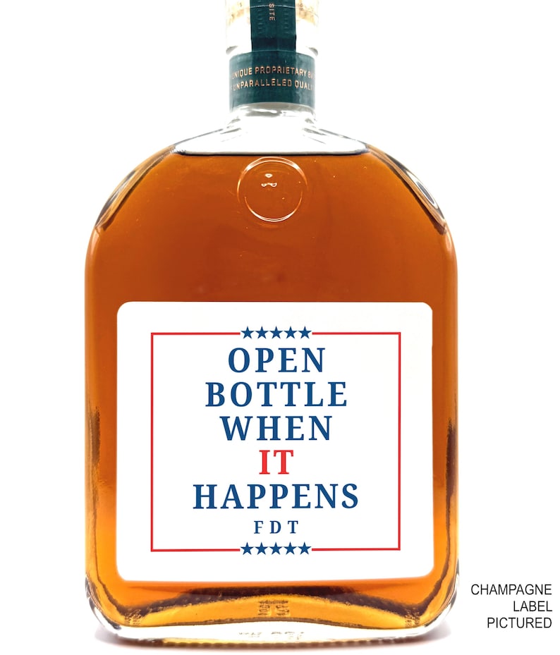 May include: A clear, square-shaped bottle filled with amber liquid. The bottle has a white label with the text "OPEN BOTTLE WHEN IT HAPPENS" in blue and red, with stars at the top and bottom. The words "CHAMPAGNE LABEL PICTURED" are at the bottom right.