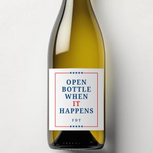May include: A wine bottle with a white label featuring the text "OPEN BOTTLE WHEN IT HAPPENS" in blue and red. The label is framed with a red border and star accents. The bottle is filled with a light-colored liquid.