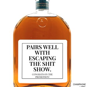 May include: A clear glass bottle filled with amber liquid featuring a white rectangular label with black text that reads: PAIRS WELL WITH ESCAPING THE SHIT SHOW. CONGRATS ON THE PROMOTION! The bottle has a dark green neck label and a circular embossed detail.