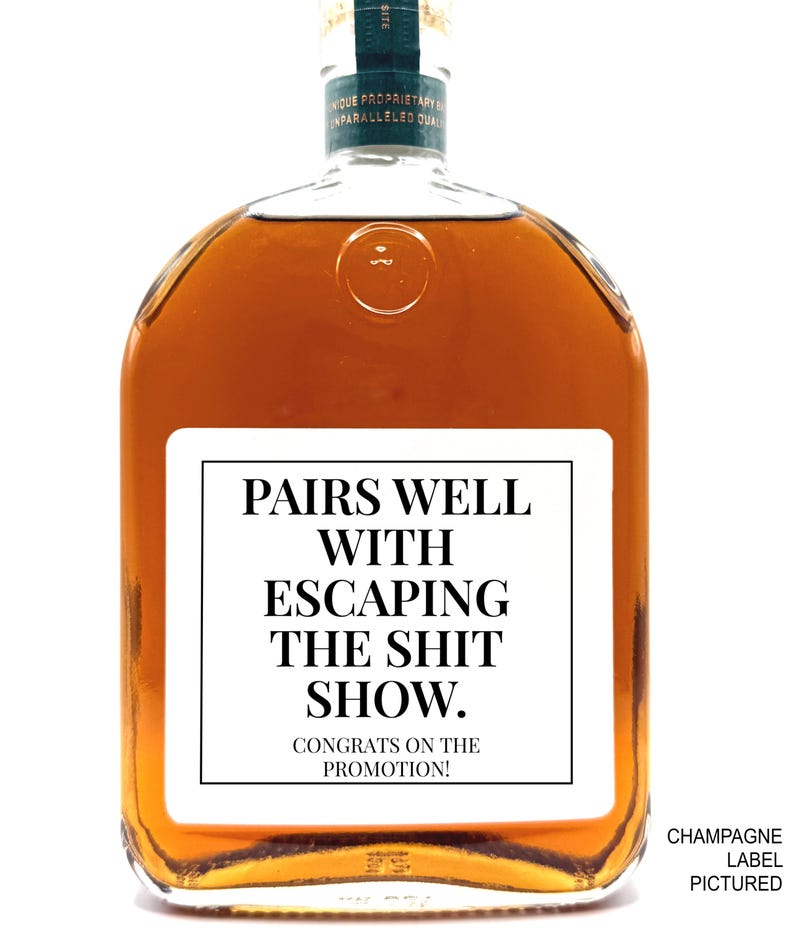May include: A clear glass bottle filled with amber liquid featuring a white rectangular label with black text that reads: PAIRS WELL WITH ESCAPING THE SHIT SHOW. CONGRATS ON THE PROMOTION! The bottle has a dark green neck label and a circular embossed detail.