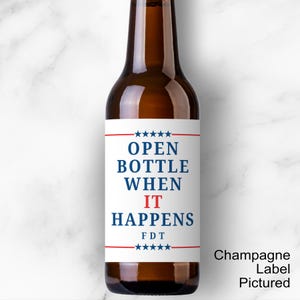 May include: A brown glass bottle with a white label. The label reads "OPEN BOTTLE WHEN IT HAPPENS" in blue and red text, with stars and lines. The bottle is on a white marble surface.
