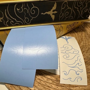 May include: Two light blue sheets of paper with a white, embossed design of stylised clouds. A white sheet of paper with a blue, stylised dragon and clouds design. The text "FOURTH WING" is visible on the top of the white sheet.