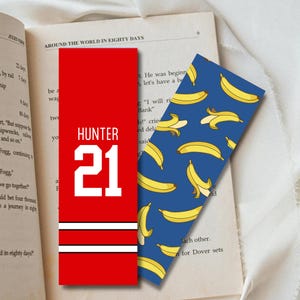 Puede incluir: Dos marcadores de libros descansan sobre un libro abierto. Uno es rojo con rayas blancas y el nombre "HUNTER" y el número "21". El otro es azul con un patrón de plátanos amarillos.