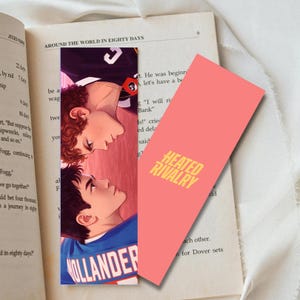 Op de afbeelding: Twee bladwijzers worden getoond met een boek. Eén bladwijzer toont een illustratie van twee mannen die naar elkaar kijken. De andere bladwijzer is koraalroze met de tekst "HEATED RIVALRY" in geel. Het boek is open op een pagina met tekst.