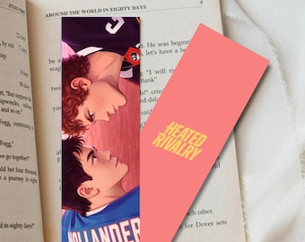 May include: Two bookmarks are displayed with a book. One bookmark features an illustration of two men looking at each other. The other bookmark is coral pink with the text "HEATED RIVALRY" in yellow. The book is open to a page with text.