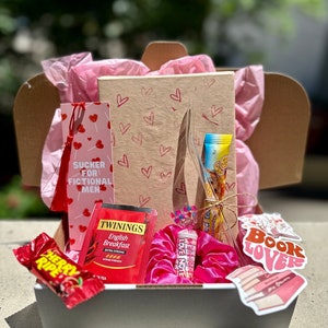 May include: A gift box filled with various items, including a pink and red bookmark with the text "SUCKER FOR FICTIONAL MEN", a pink hair scrunchie, a tea bag, a candy bar, and a sticker that says "Book Lovers".