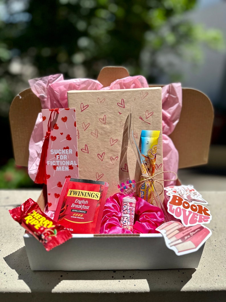 May include: A gift box filled with various items, including a pink and red bookmark with the text "SUCKER FOR FICTIONAL MEN", a pink hair scrunchie, a tea bag, a candy bar, and a sticker that says "Book Lovers".