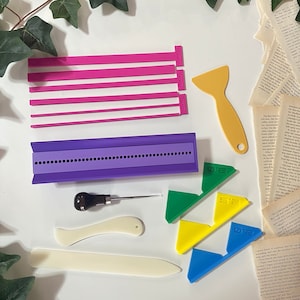 Puede incluir: Un conjunto de herramientas de encuadernación que incluye una prensa de encuadernación de color morado y blanco, una carpeta de hueso, un punzón, un raspador amarillo, un juego de cintas de lomo de color rosa y tres herramientas de esquina de color verde, amarillo y azul.