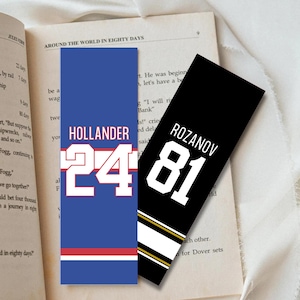 May include: Two rectangular bookmarks rest on an open book. One is blue with the name "HOLLANDER" and the number "24" in white. The other is black with "ROZANOV" and "81" in white, with gold and white stripes.