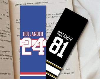May include: Two rectangular bookmarks rest on an open book. One is blue with the name "HOLLANDER" and the number "24" in white. The other is black with "ROZANOV" and "81" in white, with gold and white stripes.